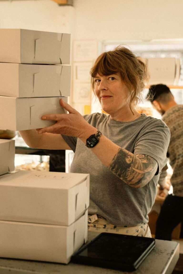 Smiling woman in a bakery arranging cake boxes with tattoos visible.