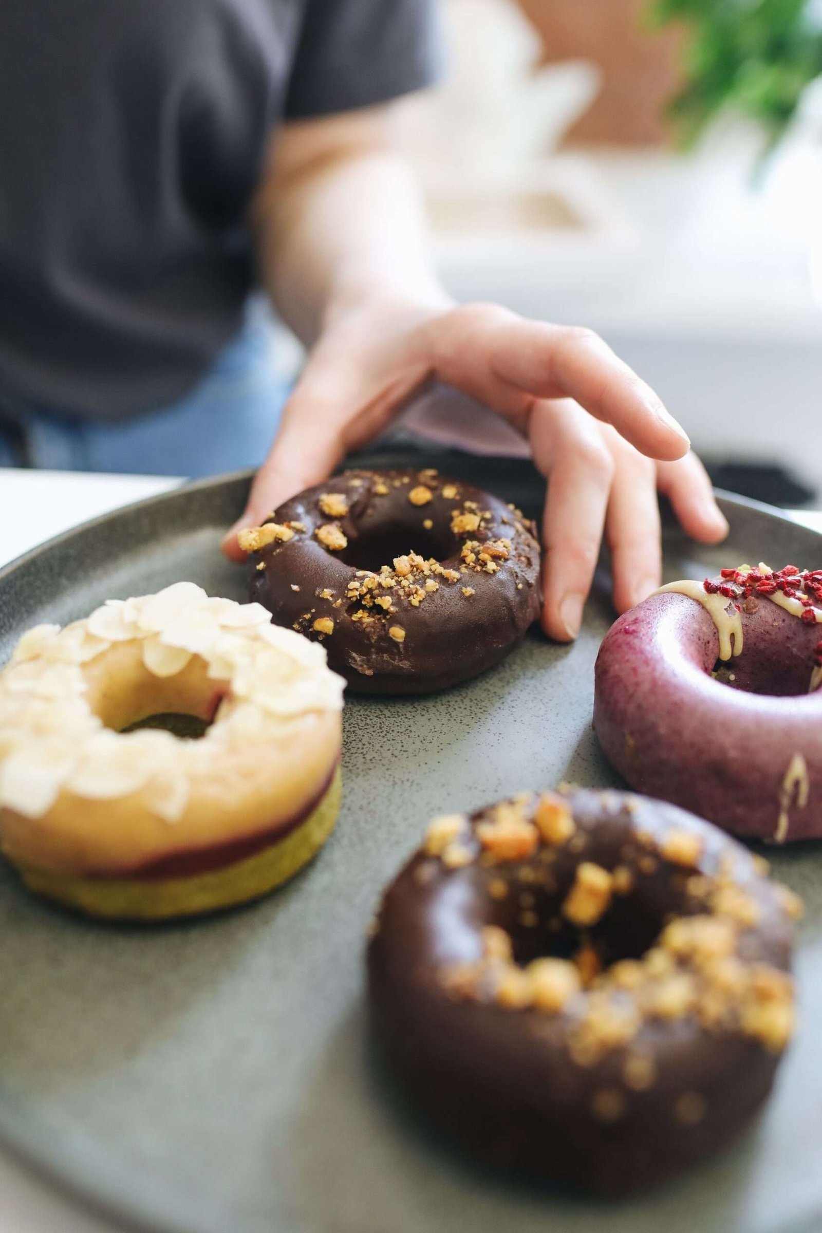 Hand reaching for an assortment of gourmet donuts on a gray plate, emphasizing variety.